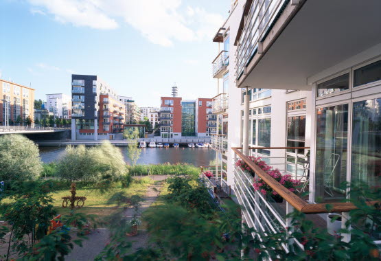 Soliga balkonger vid vattnet med blommor, utem&ouml;bler och h&ouml;ghus, vatten med b&aring;tar i bakgrunden.