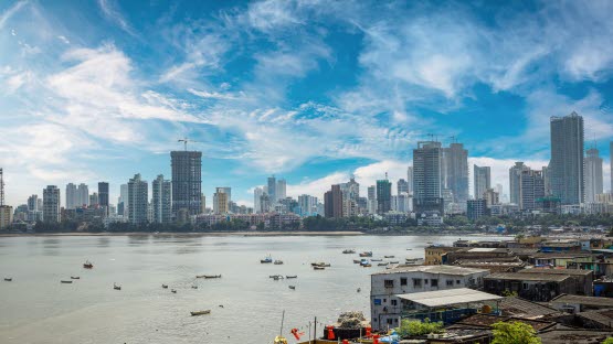 Several skyscrapers located by a bay. In front of them are several small houses. In the bay there are small boats. The sky is blue.