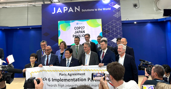 Fjorton personer står på en scen och ler. De håller upp en banderoll med texten: Paris Agreement Article 6 Implementation partnership. Bakom dem finns det en skärm där texten COP27 Japan Pavilion syns. Framför dem är en liten grupp människor som fotar. 